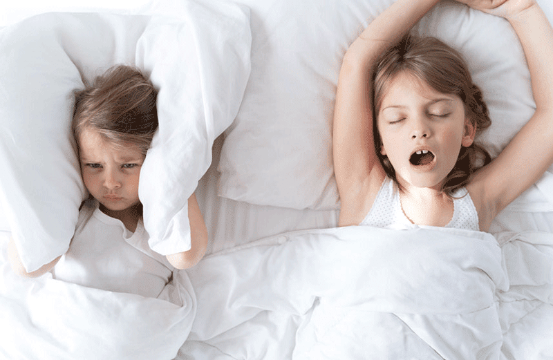 A child with a breathing disorder sleeping with her mouth open next to her sister who is covering her ears with a pillow