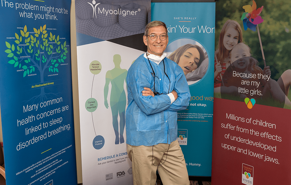 Dr. Phillips smiling in front of several posters with information about sleep disordered breathing and myofunctional therapy